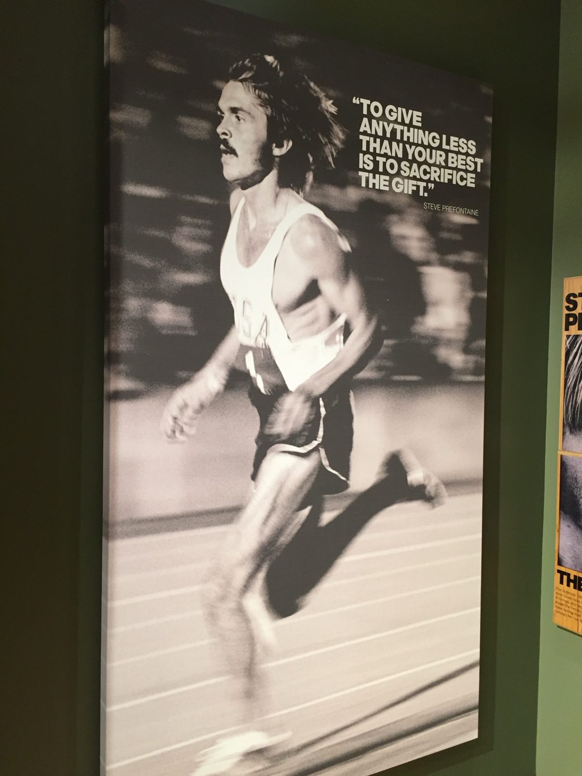 A tall canvas print at the exhibit of Pre running in a USA singlet beside his words: 'To give anything less than your best is to sacrifice the gift.'